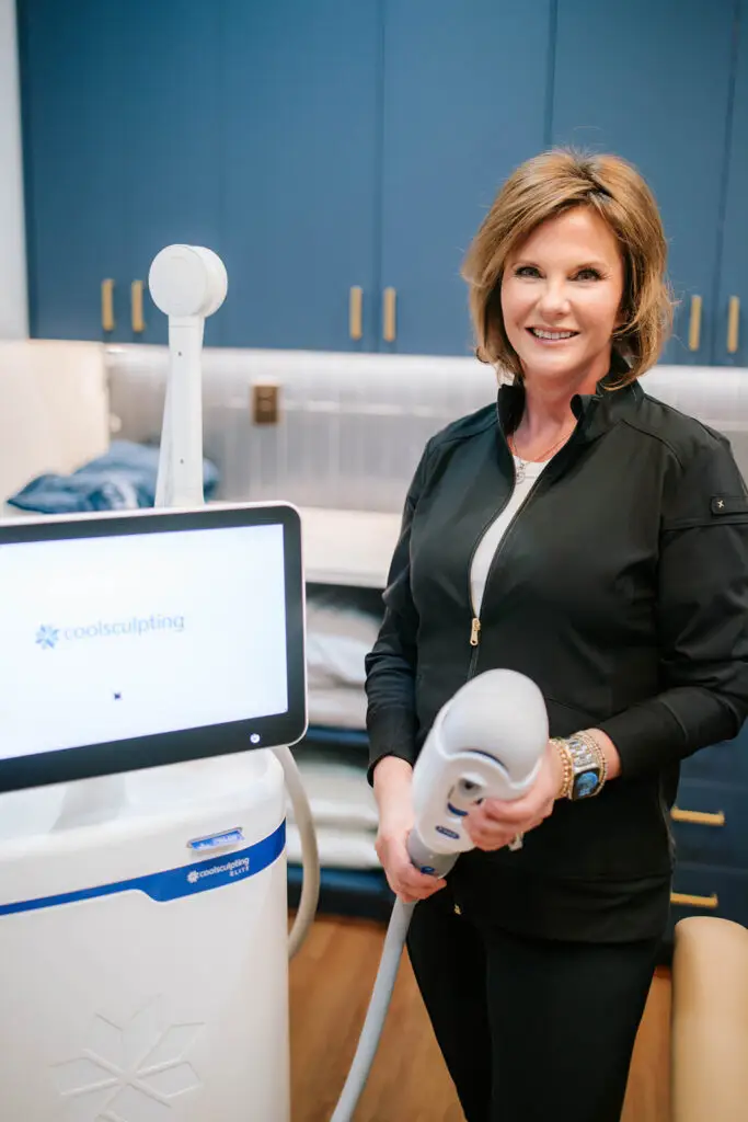 Smiling woman at Blue Spiral Medspa holding CoolSculpting Elite device in a treatment room.